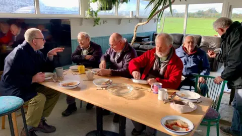 Simon Rouse Six men are sitting around a table, talking to each other. They are eating from bowls and plates full of food. There are plants and chairs in the background, as well as a green field out of the window.