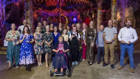 Alistair Beavis/BBC A group of people standing and one person sitting in a wheelchair. A number are holding their awards made of glass while smiling for the camera.