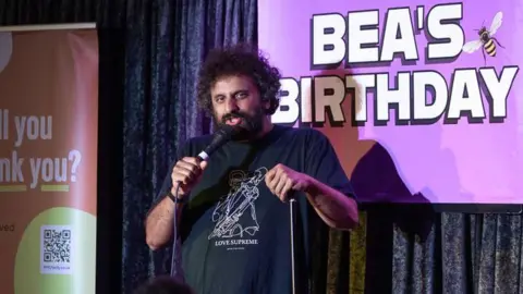 Nish Kumar on stage during the comedy event. A banner reading 'Bea's Birthday' is displayed behind him.