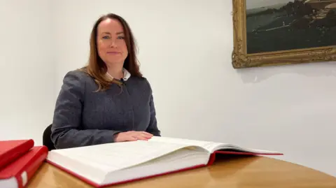 Claire is seated at a wooden table indoors with an open large ledger book in front of them and two additional red-covered books stacked nearby. A framed painting of a coastal scene hangs on the white wall behind them.