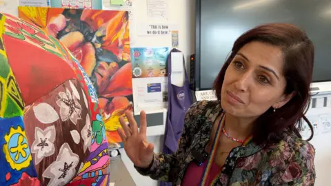 A woman in an embroidered jacket gestures towards a patchworkof colourful designs decorating a fibreglass elephant.