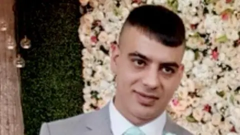 A man with short brown hair wearing a grey suit stands against a backdrop of white flowers