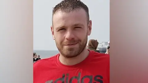 Family handout Head and shoulders of Paul Scott at a beach, the sea is in the background. Mr Scott has short, dark hair and stubble, is wearing a red t-shirt, and is smiling for the camera. 