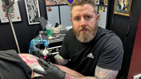 James Conway is a bearded man wearing a black T-shirt who is tattooing someone's arm at a tattoo studio. He is also tattooed and his studio can be seen behind him.