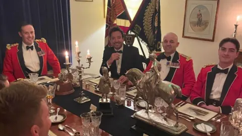 PA Media Five men sat around a dinner table with ornate decorations on it. Four of the men are wearing red blazers with bow ties and some are wearing medals.