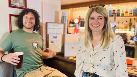 A man in a green T-shirt sits in a pub holding a pint next to a woman in a white top. Both are smiling at the camera.
