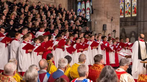 Dale Hodgetts A large choir performing inside Gloucester Cathedral as part of the opening service for the Three Choirs festival. A number of young choristers at the front are wearing white and red gowns, with adult performers behind them dressed in black