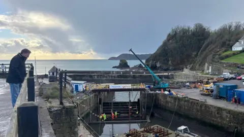 BBC Charlestown Harbour lock gate. There is a crane over the area and men in high vis jackets work down on the water.
