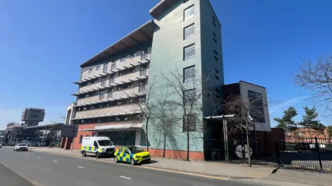 Two police vehicles are parked outside the former YMCA building in Liverpool on a sunny day. The building is a large block with six floors.