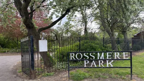 A metal sign reads Rossmere Park in white letters inside a black frame. A black metal fence with an open gate leading into the park can be seen beyond. Trees and thick foliage borders the path into the park.