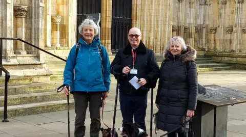 Clare Slator Three people stand in front of a grand Gothic cathedral entrance, dressed in winter clothing. Two women hold leashes attached to dogs, and one holds a piece of paper. The cathedral features pointed arches, intricate stone carvings, and a large circular stained glass window above the doorway.