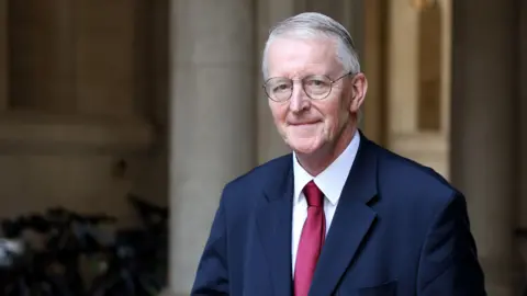 Getty Images Hilary Benn arrives at 10 Downing Street. He's wearing glasses and a navy suit with a white shirt and a red tie. 
