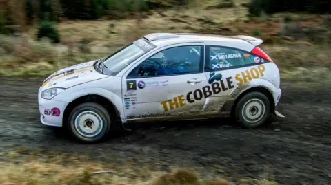 Rally Media UK A white rally race car travels at speed on a gravel track. The car has sponsorship stickers plastered across the side of the car, one reading 'THE COBBLE SHOP'. The vehicle is travelling in a forest. 