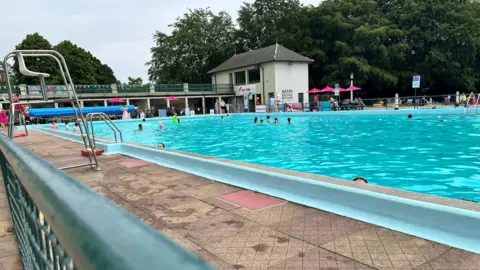 Emma Baugh/BBC The swimming pool has several people swimming in it. In the background there are umbrellas and seating. On the left in the foreground is a white lifeguard chair, which is unoccupied.