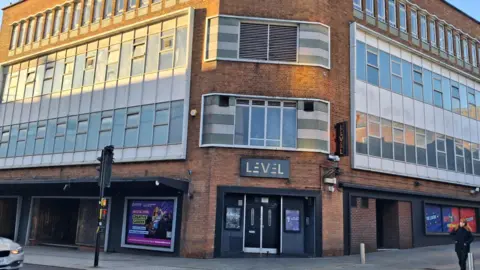 It is a three storey building with grey panelling around the rows of windows. The building stands on a street corner with two black doors visible in the centre.