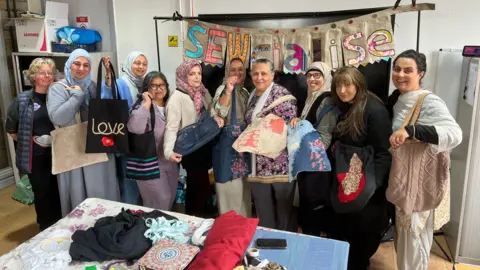A group of ladies stand in front of a big sign which says 'Sewcialise'. The sign is made of handmade textile bunting, and the women are all holding up tote bags made of different materials. In front of them is a table full of rolls and piles of colourful textiles, boxes of thread and other related items.