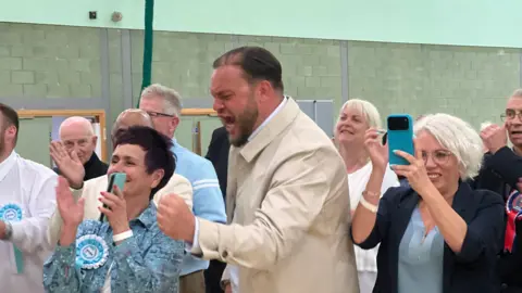 Simon Dedman/BBC Sam Journet, a Reform councillor wearing a cream jacket, appears to shout and clench his fist in a sports hall, surrounded by other people clapping.