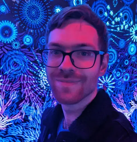 James Dyer A man with short dark hair and dark-rimmed glasses is smiling at the camera, visible from the shoulders up. He is in a pink-tinted light and behind him is a blue background with lots of lights and patterns.