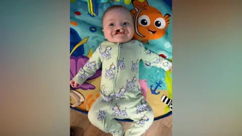 A baby, Eli. He is lying on a colourful playmat that has an underwater design, including fish, anchors and seaweed. He is wearing a green baby grow with Zebras.