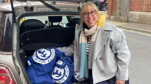 Lisa Pike wearing a grey woollen coat with a blue, cream, yellow and navy scarf. She has blonde hair and black glasses. She is stood by the boot of a car with resting on several blue tote bags.