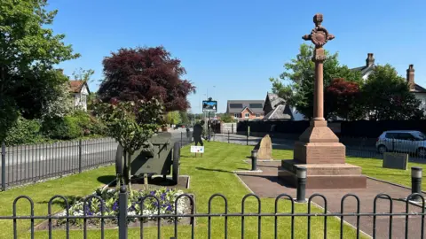 Hale Village cenotaph in a fenced area in the middle of Hale village. A sandstone war memorial is surrounded by grass and flower beds.