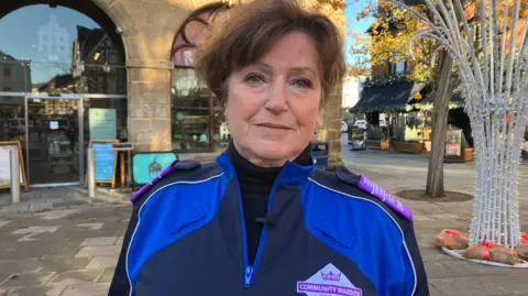 BBC A woman with dark hair and wearing a blue top with two different shades of blue is looking at the camera. The top includes a purple and white badge including the words community warden. 