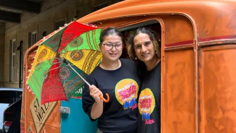 Two women look out of a brown caravan. The woman of the left is holding a colourful umbrella