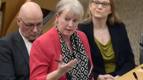 Shona Robison, who has blonde hair tied back, speaks in the Scottish Parliament chamber with her right hand held out in front of her, holding a pen. She is wearing a pink suit jacket and a black and white scarf. 