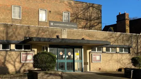 The front of Harpenden Public Halls, which is a two-storey building built out of cream coloured bricks. There are three windows on the top floor, which has a flat roof. The entrance doors are green with windows on them and they have notice boards either side. There are two green shrubs outside the entrance.