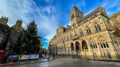 The exterior of Northampton Guildhall set against a blue sky with patches of cloud. A Christmas tree is visible opposite the building.