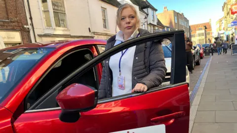 Andrew Sinclair/BBC Care provider Katie Thorogood standing next to a car with "Love Life Care" written on the door.