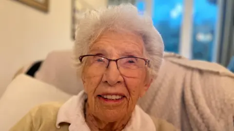 BBC Pat Shepherd has white hair and glasses. She sits on a beige sofa and smiles at the camera.