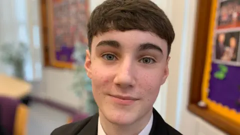 James has short brown hair, is wearing a black blazer and a purple and black tie. The background is a school setting with work on the walls.