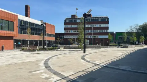 A paved open space, with trees and lighting columns. The space is bordered by tall buildings. There are benches and planting located around the paved area.