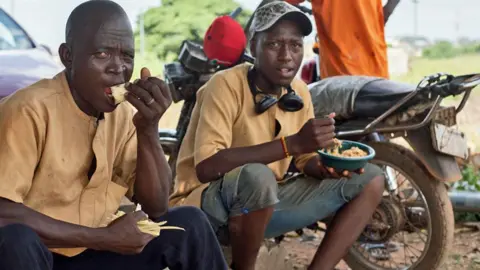 Ifiokabasi Etang/BBC Two cemetery workers sit under a tree eating at lunchtime – a motorcycle can be seen behind them