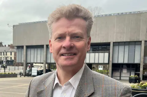 BBC/David Macmillan Matthew Snedker has short grey hair, brown chequered formal jacket and white shirt. He is standing in front of a grey building.