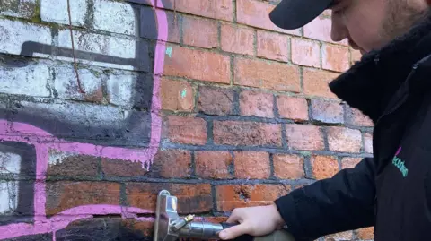 A red brick wall with pink and white graffiti on it is being cleaned using an industrial cleaner by a man wearing a cap and fleece. 