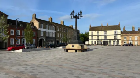 Tom Jackson/BBC A completed Market Square on a sunny day with blue skies. The ground has been paved in dark and light grey slabs, with some red patches. In the centre is a large black street lamp with seating around it. There are bollards at the edges of the square.