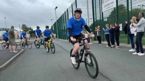 Five boys in blue tshirts and cycle helmets on bicycles with a number of adults on either side of them clapping and a green-fenced astroturf area behind them
