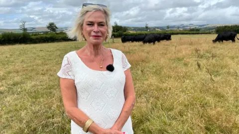 Rachel Gilmour is standing in a field with cows grazing in the background. She is wearing a white dress and she has blonde hair in a bob. She has a pair of sunglasses on her head.