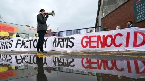 PA Media A protester holding a megaphone outside BAE Systems in Glasgow in May 2024. Behind him are red brick walls and the gates of the premises, and a long banner which reads "This factory arms genocide". The image is reflected in a puddle of water.