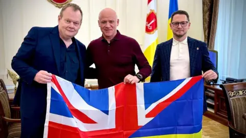 Cllr Paul Goggin (left) with Mayor Gennadiy Trukhanov (centre) and Deputy Mayor Pavlo Vugelman (right) holding the flags of the United Kingdom and Ukraine.   