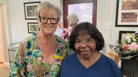 Two women look straight ahead smiling. The woman on the left wears a bright floral dress and distinctive glasses, and the woman on the right wears nurses scrubs.