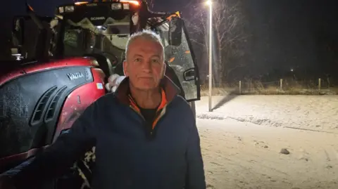 Duncan Wight A man with white hair wearing a blue fleece stands in front of a snow plow and a snowy road.