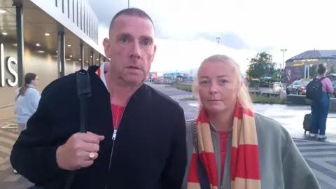 A man with short dark hair wearing a black jacket at red Liverpool jersey underneath. A woman is on the right, with blonde hair tied back, wearing a red and cream scarf and grey jacket.