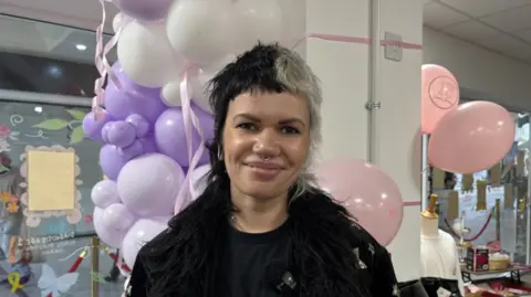 A woman with black and white short hair looking into the camera, her hair is split into the two colours. She is smiling and has a septum piercing. There are purple and pink balloons behind her. 