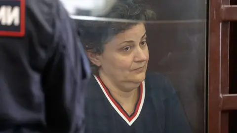 BBC Anna sitting behind the glass screen of the court cage with a wooden frame. She is on the right side of the shot, looking towards the right with a serious expression on her face. She has short dark hair, wearing a navy v neck jumper with red trim. On the left-hand is the back of a security guard with Russian writing on his navy uniform.