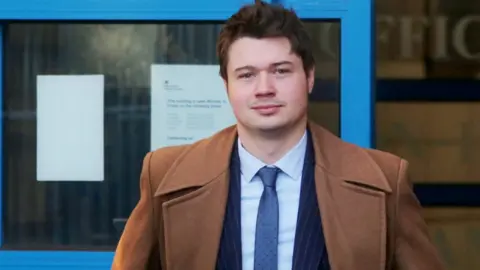 SWNS A man with short dark hair stands outside the door of a building with blue window frames. He is wearing a dark blue pinstripe suit and brown overcoat.