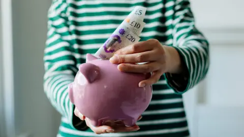 Woman putting money into a pink piggy bank. She is wearing a green and white striped top.