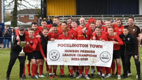 Pocklington Town Football Club The team stand in red socks and jerseys and two players are holding up the silver cup. Others hold a sign that reads Pocklington Town, Humber Premier League Champions 2022-2023.